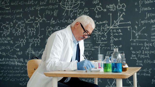 Researcher in lab coat working on a desk with glassware and chemicals, with a large blackboard in the background. 