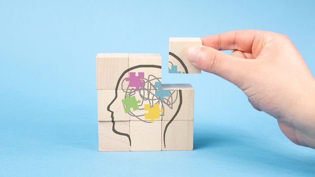 Hand placing puzzle block into wooden head silhouette with colorful brain pieces on blue background. 