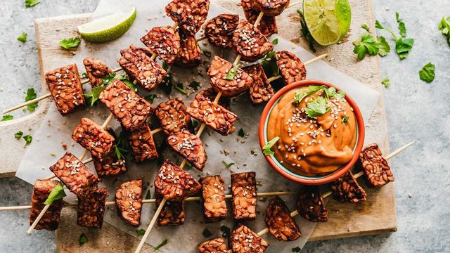 Grilled tempeh protein skewers topped with sesame seeds, served with creamy peanut sauce and lime wedges. 