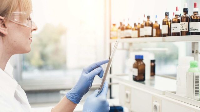 Researcher in lab wearing gloves uses a tablet beside shelves of chemical bottles. 