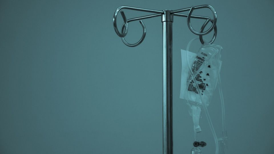 Intravenous fluid bag hooked onto a metal hospital stand, against a gray background.