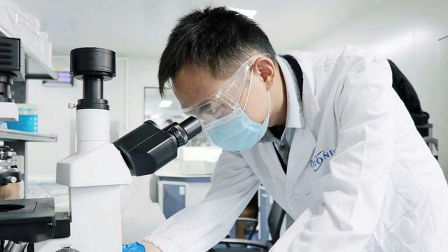Researcher in laboratory wearing a lab coat and goggles, examining samples under a microscope. 