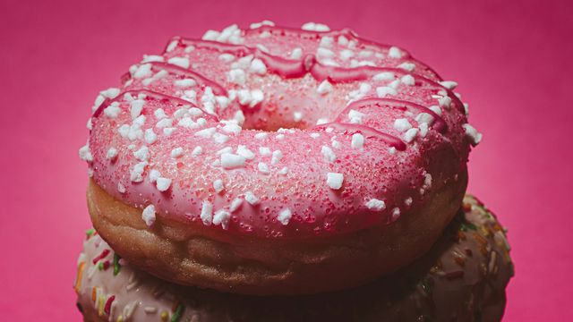Pink frosted donut with sprinkles and sugar crystals against a bright pink background. 
