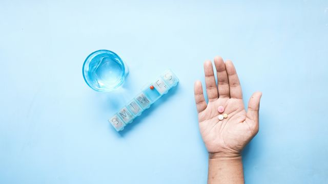 Birds-eye view of medication within the palm of a hand, next to a glass of water and dossette box, on a blue background. 