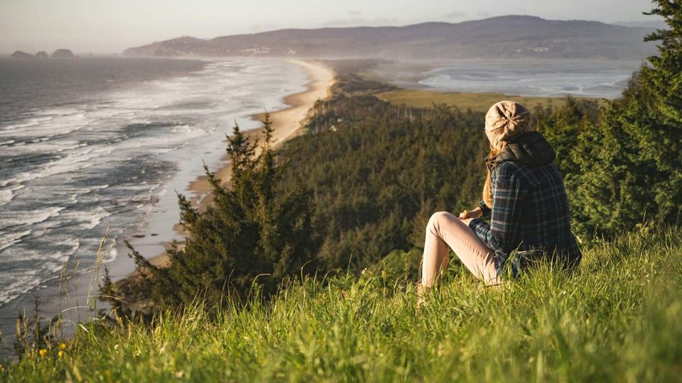 Person sitting on grassy cliff overlooking the ocean and sandy coastline at sunset.