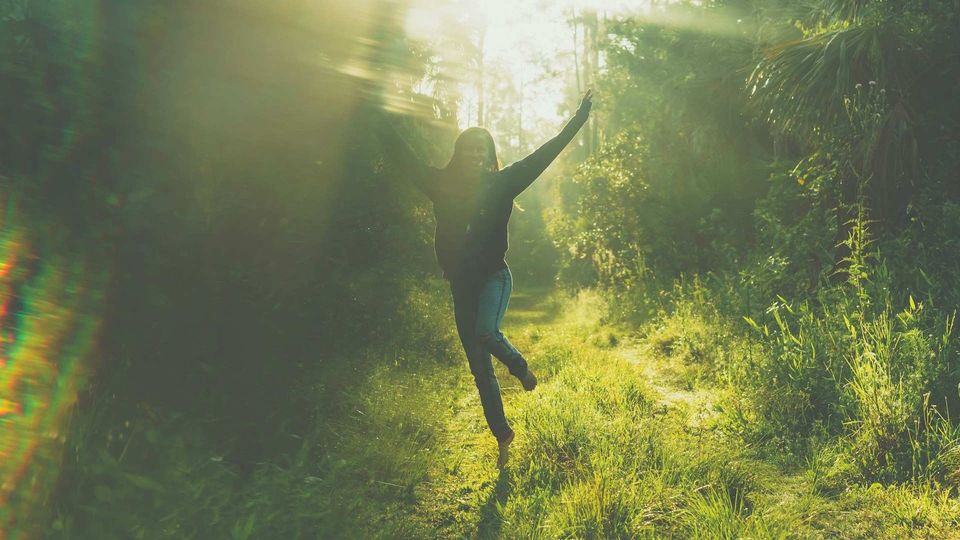 Woman jumping with arms raised on a sunlit forest path surrounded by trees.