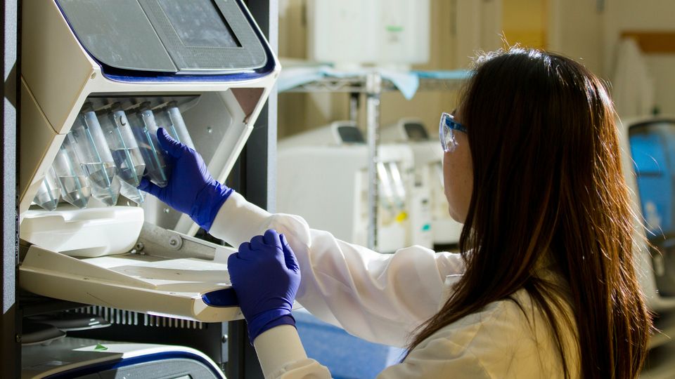 Researcher in laboratory setting, placing test tubes into a machine while wearing protective gloves and goggles.