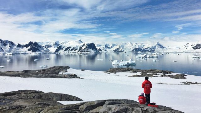Two people in red jackets standing on the edge of a snowy and rocky Antarctic coastline. Across the water, glaciers are visible. 