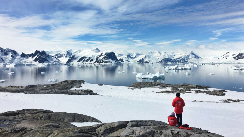 Two people in red jackets standing on the edge of a snowy and rocky Antarctic coastline. Across the water, glaciers are visible.