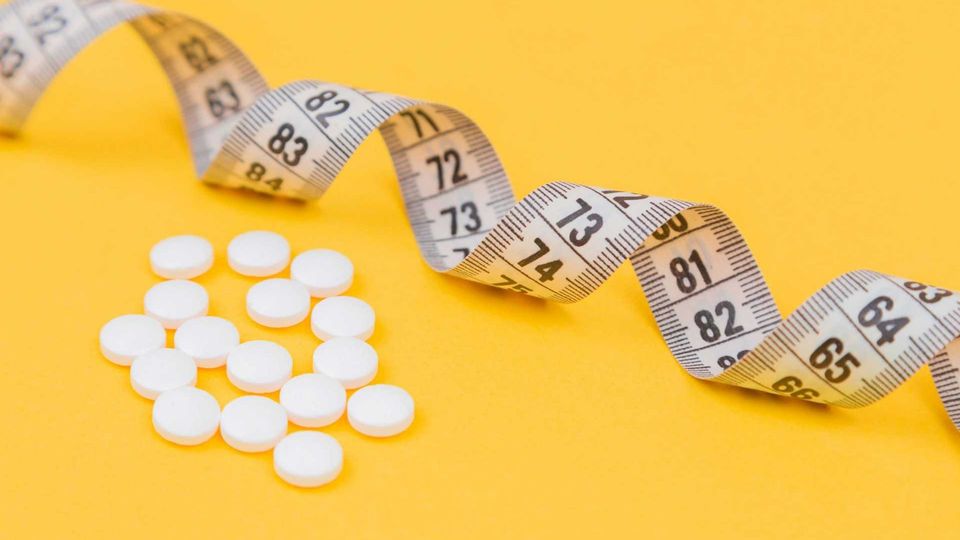 Multiple white pills beside a rolled out measuring tape, placed on yellow background