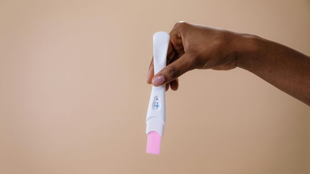 Hand of a woman holding a positive pregnancy test against a brown background. 