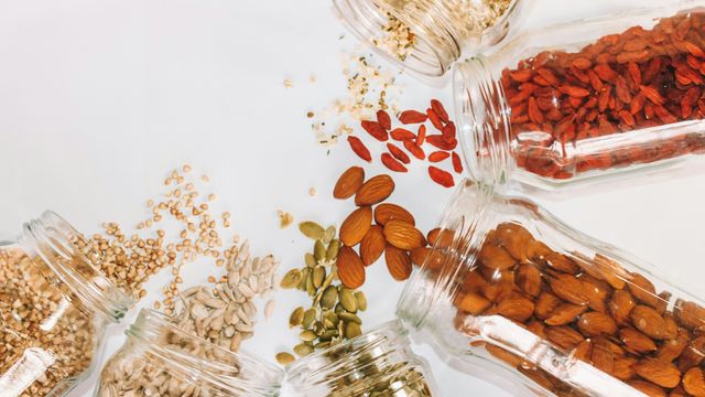 Glass jars lying horizontally on a table, with nuts, seeds, and other wholegrains spilling out onto the table. 