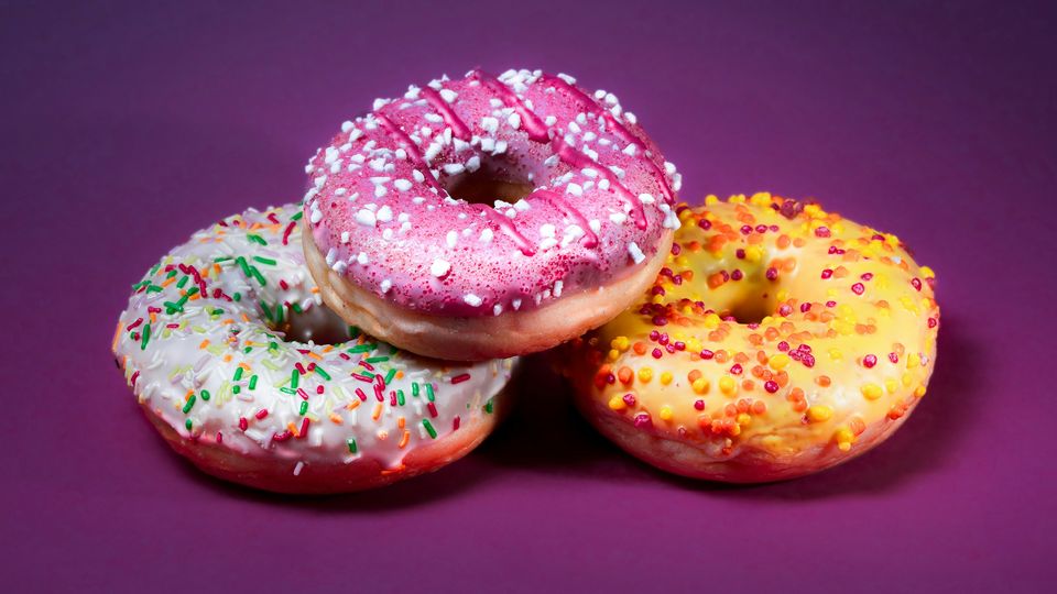 A white donut, pink donut, and a yellow donuts, set against a purple background. Each donut is topped with multicolored sprinkles.