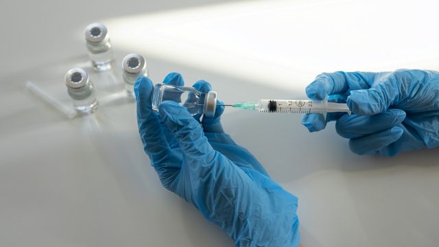 Gloved hands using a syringe to withdraw liquid from a vaccine vial, with multiple vials in the background. 