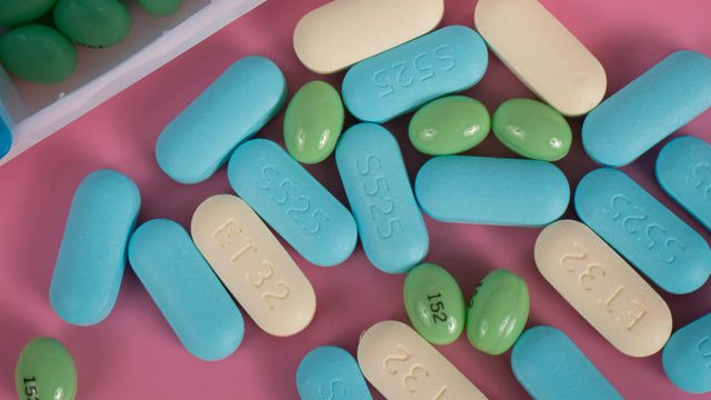 Assortment of yellow, green, and blue pills on a pink surface, next to an open pill box containing more tablets. 