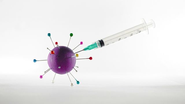 Syringe entering a virus model, represented by a purple ball with multiple pins inserted, against a white background. 