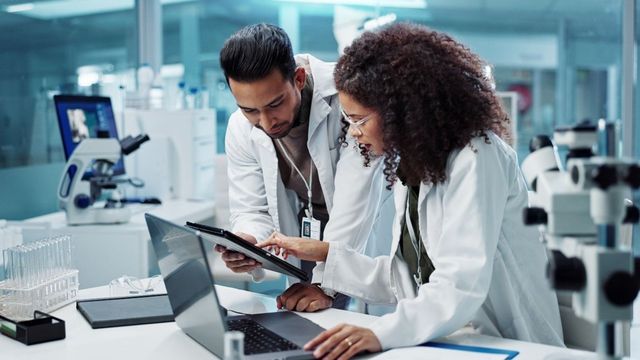 Researchers reviewing data on a tablet in a modern laboratory with microscopes and equipment. 