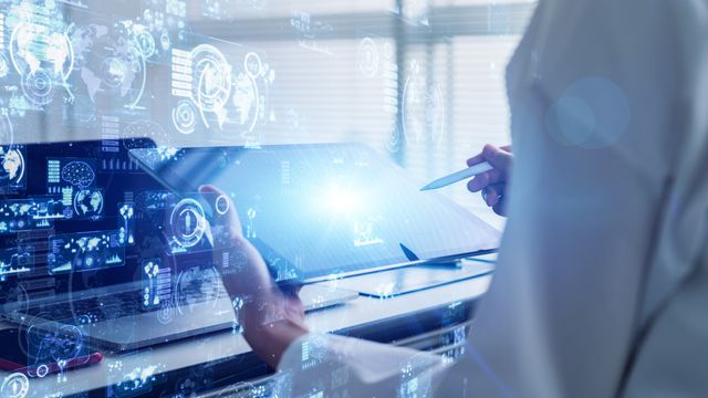 A person in a lab coat uses a touchscreen device at a workstation, with blue digital interface graphics overlaid. 
