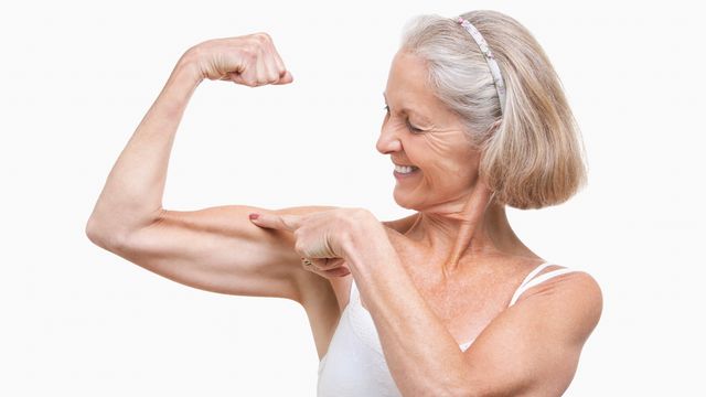 An older women flexing her muscles and pointing to her biceps against a white background. 