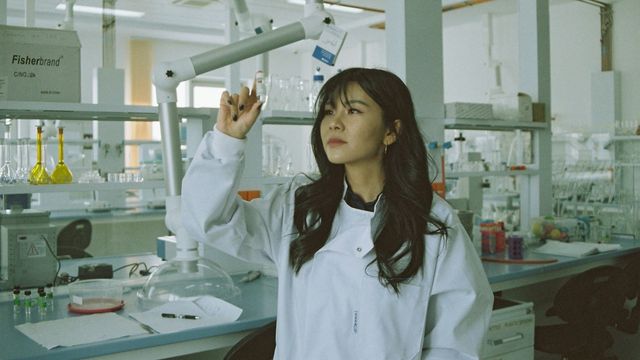 Scientist in lab coat examining a sample in a laboratory setting with glassware. 