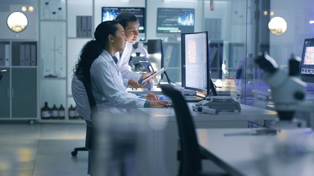 Two female scientists in lab coats looking at microscopy data on a computer in a lab. 