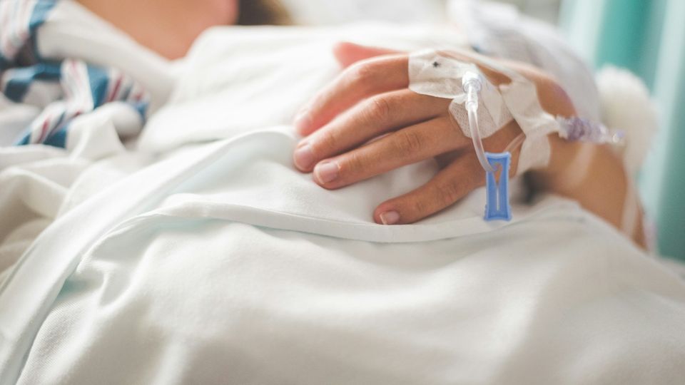 Close-up of a patients hand with an IV drip line inserted, rested on hospital sheets.