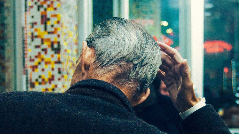 The back of an elderly man's head, as he reaches up to touch his forehead with his right hand.