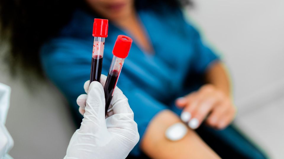 Gloved hand holding two test tubes containing blood samples. In the background, a woman is pressing a cotton ball to her inner elbow.