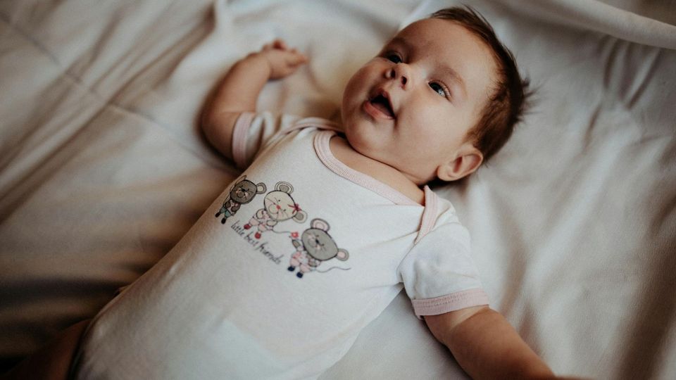 Baby lying on bed wearing a onesie, looking up with a curious expression.