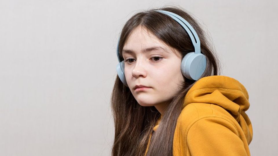 A girl with long dark hair wearing a yellow jacket and overhead earphones looks sadly toward the floor.