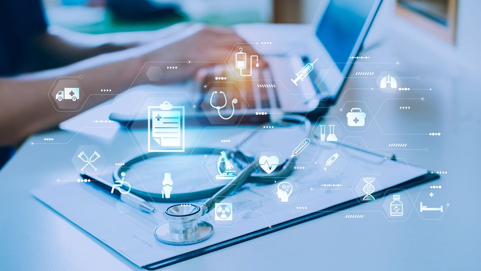 Seated doctor using a laptop. Next to the laptop is a clipboard, stethoscope and a hologram of AI and medical icons.