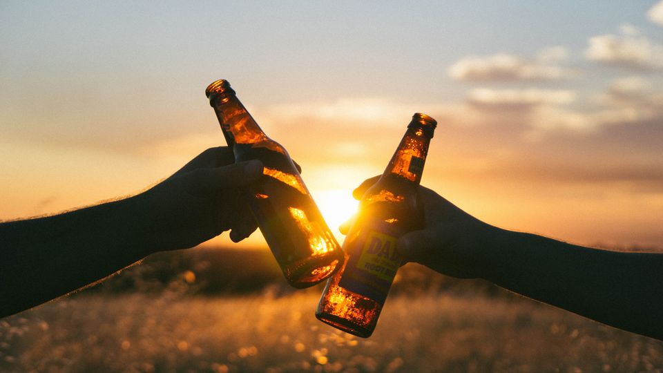 Two people clinking beer bottles at sunset, representing alcohol consumption and social drinking.