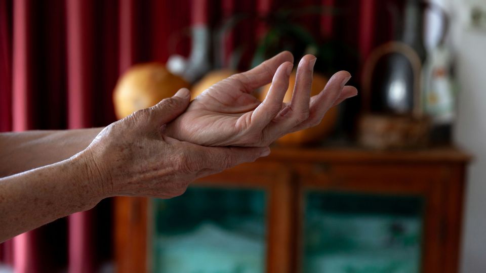 The hands of a person holding their left wrist, with a display cabinet and red curtain seen in the background.