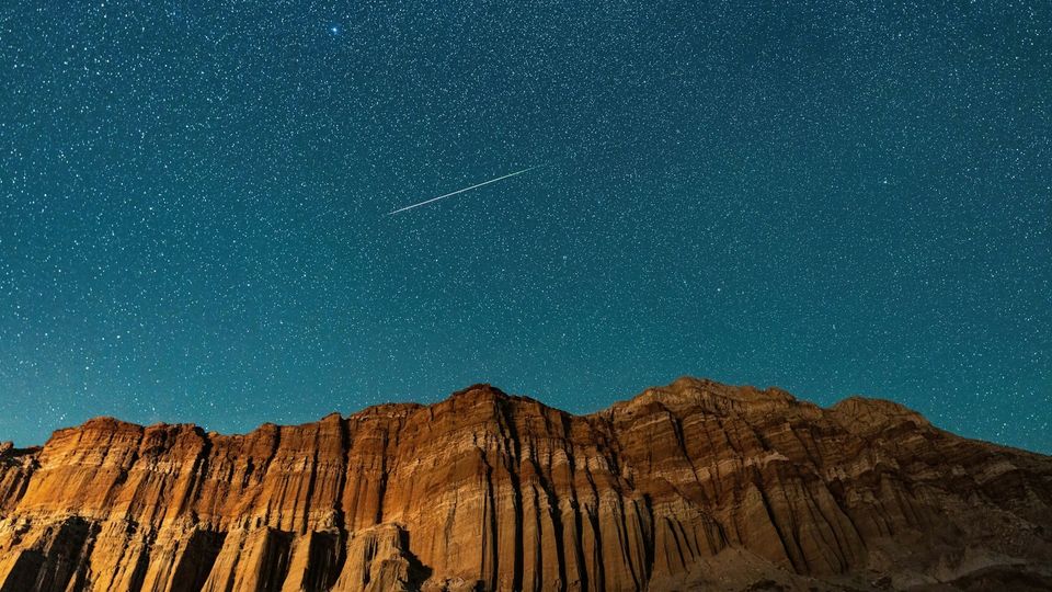 Starry night sky over rugged mountains with a shooting star streaking above the cliffs.