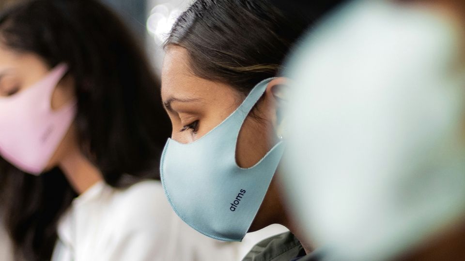 Side-view of a woman wearing a pale blue face mask, surrounded by other people wearing masks in an outdoor environment.