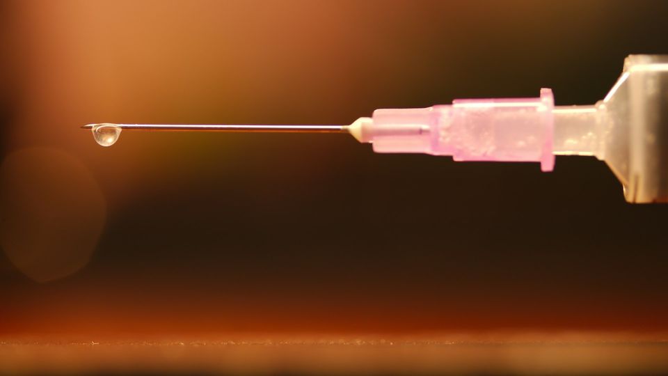 Close-up of a vaccine needle releasing a single droplet, against an orange background.