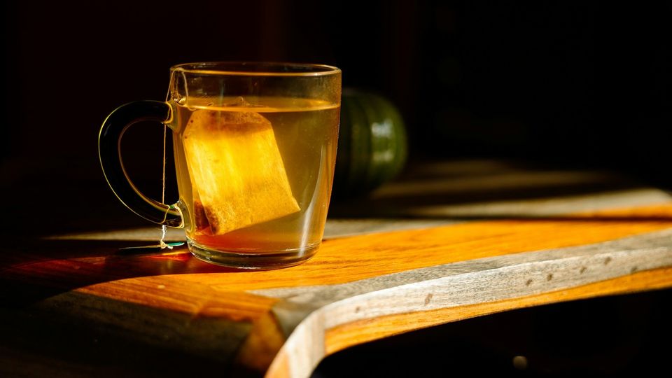 Glass teacup with a steeping tea bag on a wooden table in warm sunlight.