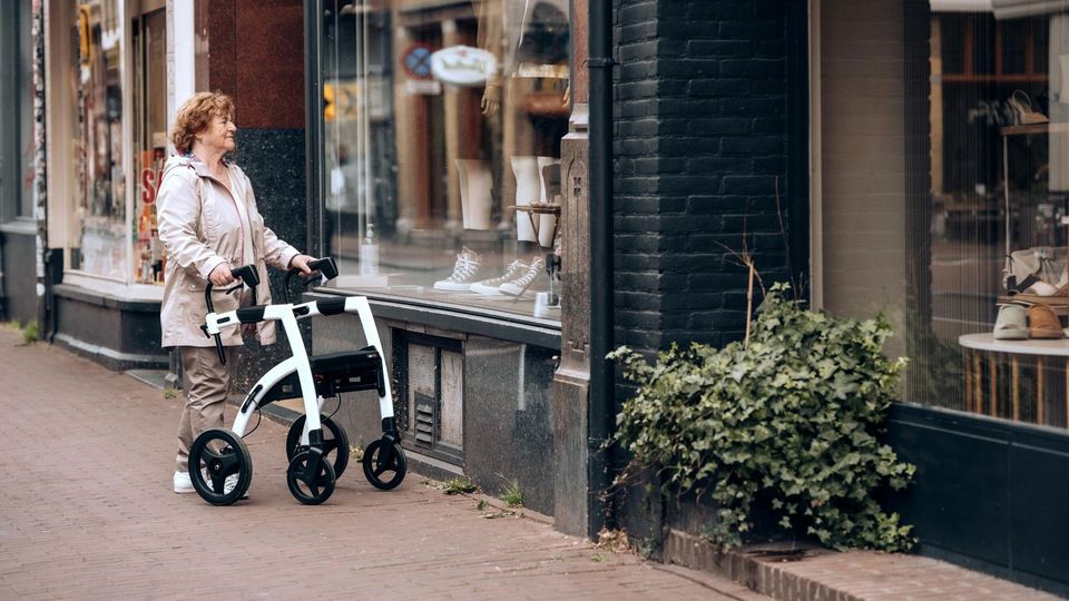 Elderly woman using a walker while looking at a shop window on a city street.