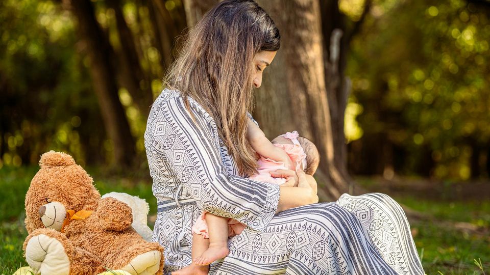 Mother breastfeeding baby outdoors in a park, with teddy bear beside them.