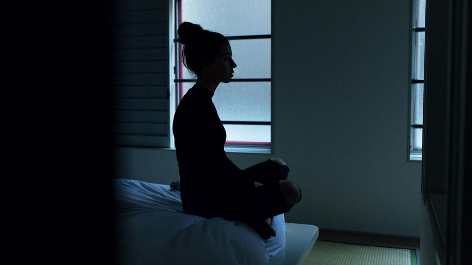 Silhouette of a woman sitting cross-legged on her bed in a dark room.