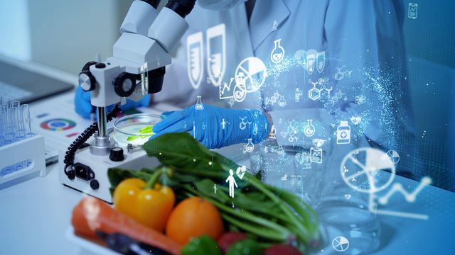 Scientist wearing a white lab coat and blue gloves examining a food sample under a microscope next to an assortment of fruit and vegetables.  