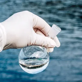 Gloved hand collecting water sample for validated EPA 1633 PFAS workflows across environmental and biological matrices.