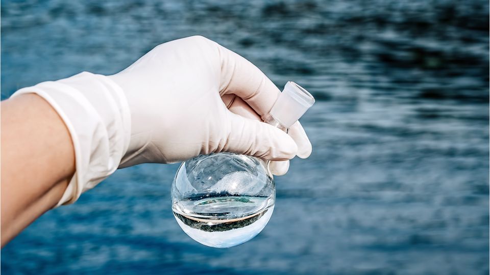Gloved hand collecting water sample for validated EPA 1633 PFAS workflows across environmental and biological matrices.