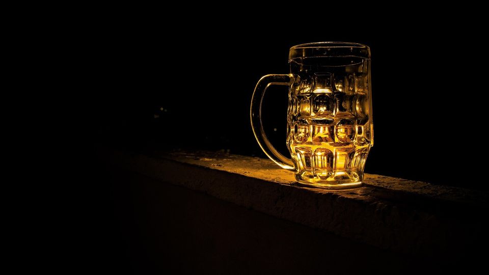 Glass beer mug filled with golden beer on ledge against dark background.