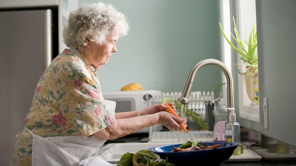 An older woman washing vegetables by a kitchen sink.