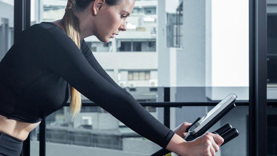 Woman exercising on a stationary bike in a modern gym with large city-view windows.