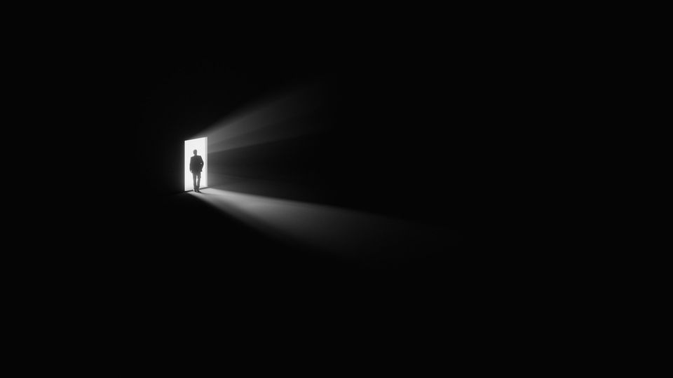 Silhouette standing in doorway with light piercing darkness, symbolizing isolation and uncertainty.