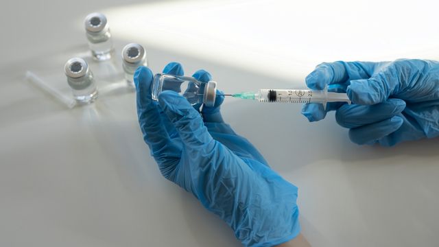 Gloved hands using a syringe to withdraw liquid from a vaccine vial, with multiple vials in the background. 
