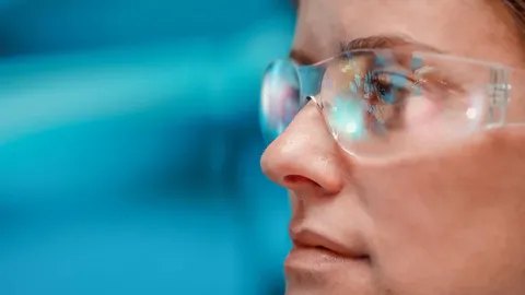 Close-up of scientist wearing safety glasses with digital data reflected, against blue background.