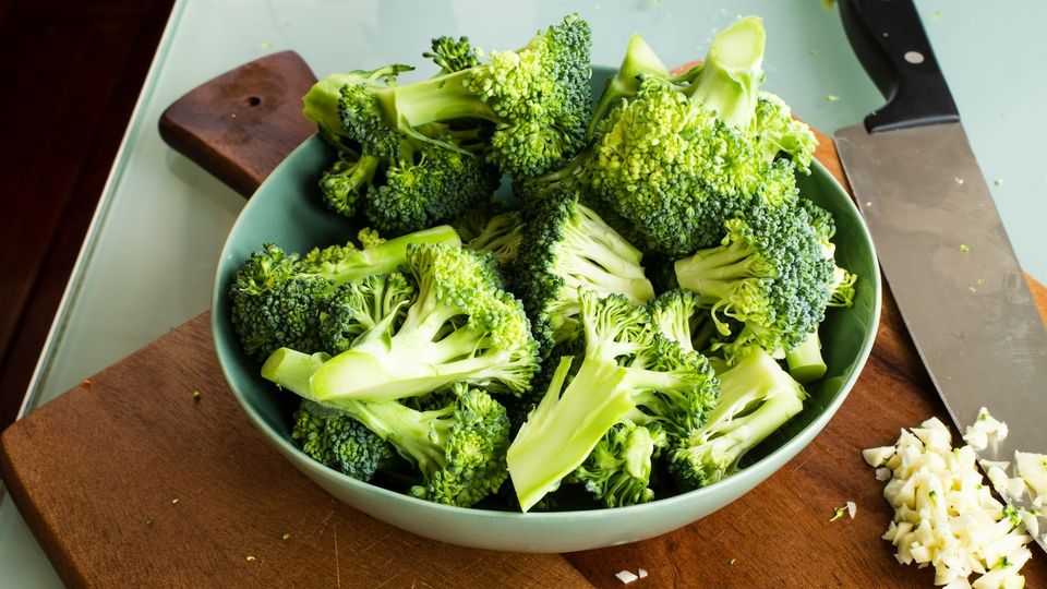Broccoli florets in a bowl on a cutting board with chopped garlic and kitchen knife.